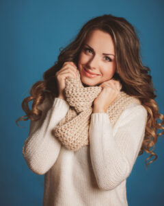 Woman with long curly brown hair wearing a sweater and scarf, holding the scarf up to her face