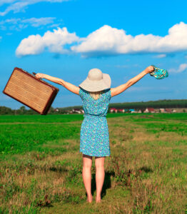 Woman wearing a dress and sun hat in a grassy field holding her sandals in one hand and luggage in the other