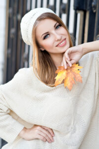 Woman in a white sweater and white knit hat holding a yellow and orange leaf