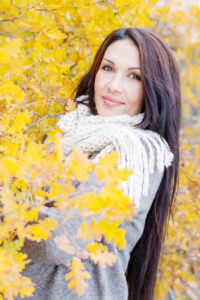 Woman with long brown hair wearing a scarf and sweater standing in a bush of yellow fall leaves