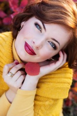 Red-haired woman in a yellow sweater smiling outdoors next to flowers