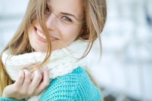 Woman in a blue knit sweater and white scarf smiling with her long brown hair blowing in the wind