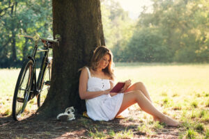 Woman in a white dress sitting against a tree reading a book with her bike propped against the tree