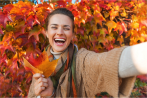 Joyful woman smiling in front of a background of fall leaves while holding a large orange leaf