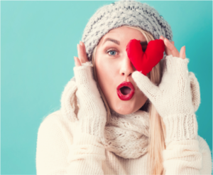 Blonde-haired woman wearing white gloves, sweater and scarf holding a plushie heart over one eye