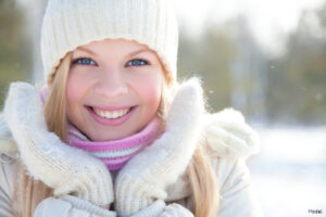 Smiling woman in a white hat, gloves and winter coat framing her face with her hands outdoors