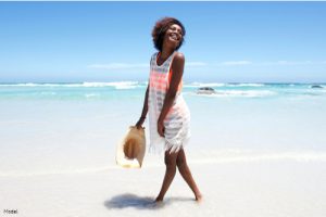 Joyful, confident woman walking along the shore of a beach in an orange bikini, semi-sheer white cover-up and holding a beach hat