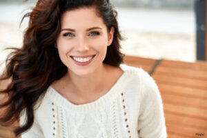 Smiling woman with clear skin and long brown hair wearing a white knit sweater