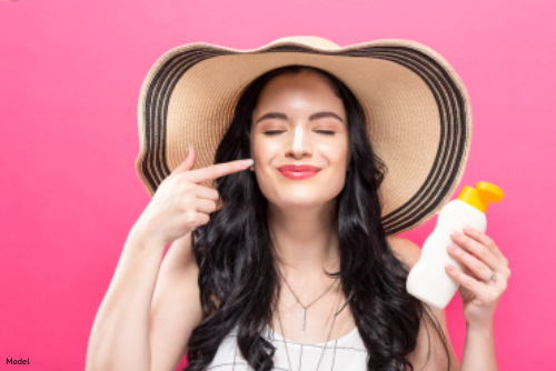 Happy woman in a large sun hat holding sunscreen and pointing to her face
