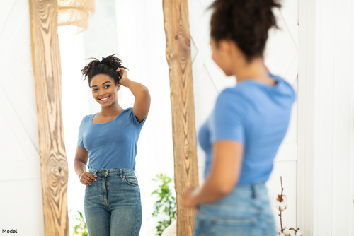 fit woman wearing blue shirt and jeans, looking into a mirror and smiling