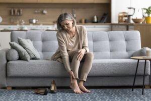 Woman sitting on a sofa after work, massaging her tired feet and touching her ankle in discomfort, highlighting symptoms of varicose veins and the need for rest, orthopedic insoles, or vein care treatments like sclerotherapy