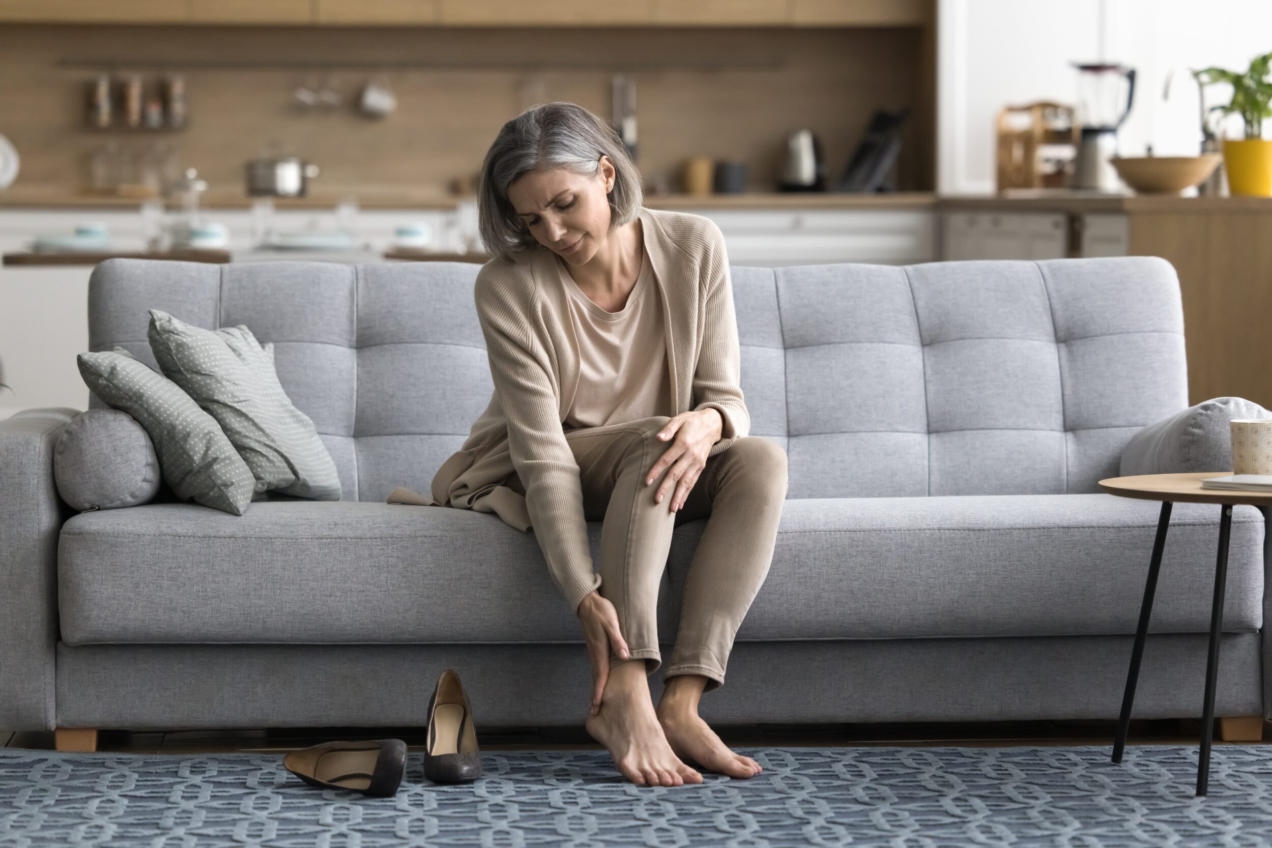 Woman sitting on a sofa after work, massaging her tired feet and touching her ankle in discomfort, highlighting symptoms of varicose veins and the need for rest, orthopedic insoles, or vein care treatments like sclerotherapy