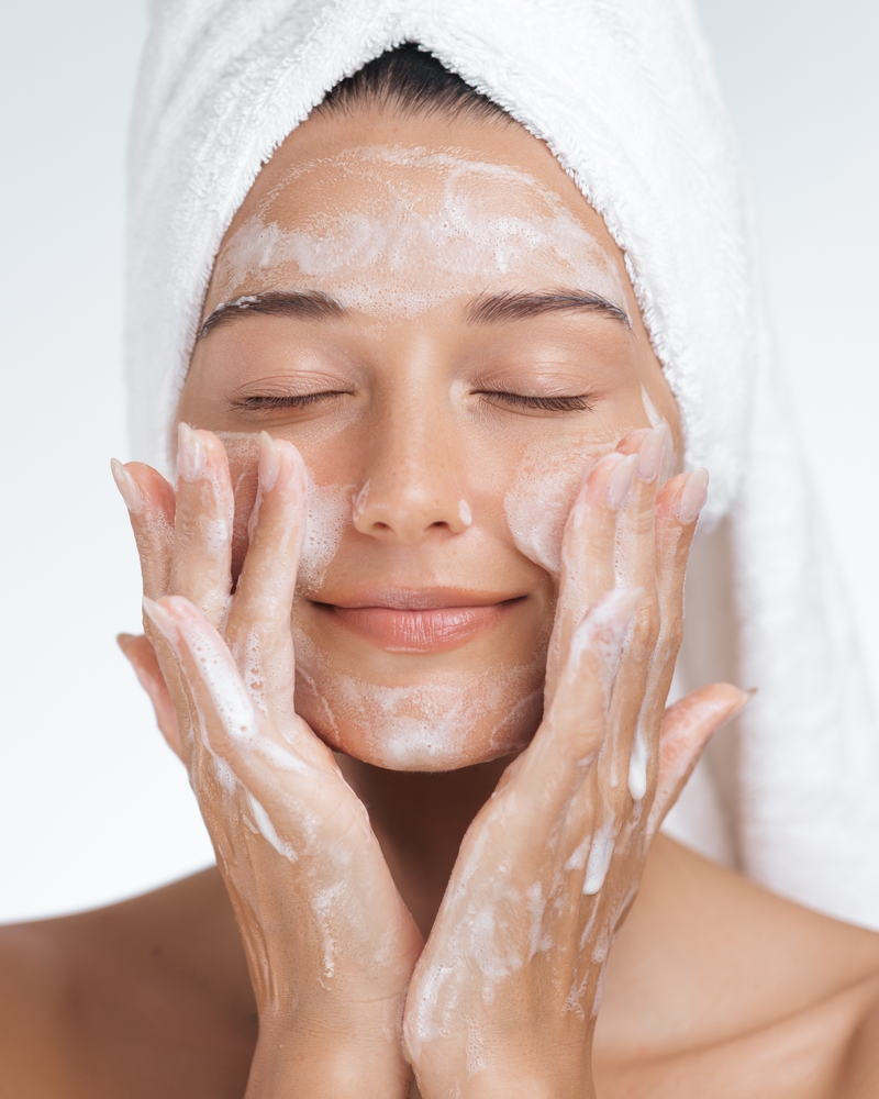 Relaxed woman with towel on head massaging foaming cleanser into skin, concept of beauty, self-care, and soothing skincare routine.